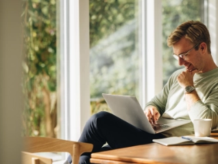  Young man in glasses and a light sweater working happily on a laptop at a wooden table in a bright home office, with large...